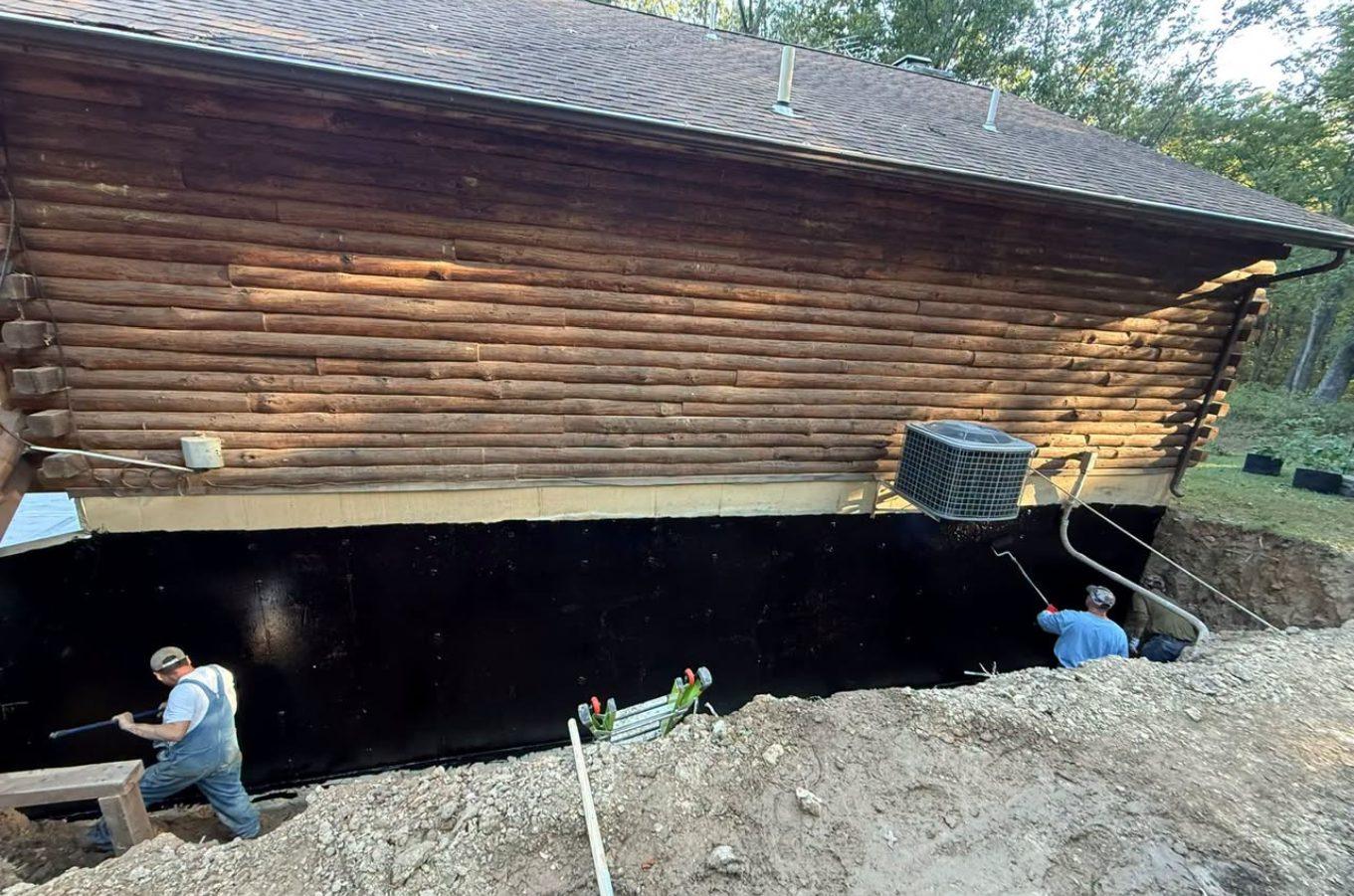 Crew applying black waterproofing to an excavated foundation wall under a log home